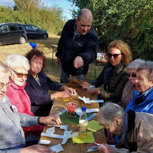 An einem Biertisch im Freien sitzen Menschen beieinander. Hinten am Tisch steht der Pastoralreferent Meier.